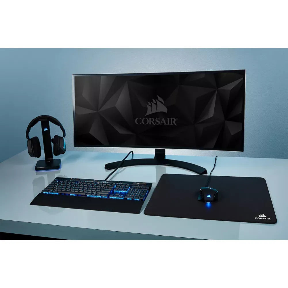 Corsair MM100 gaming mouse pad displayed with a monitor, keyboard, and headset on a modern desk.
